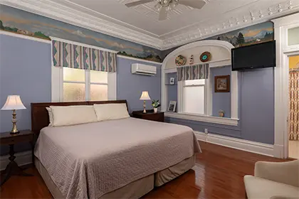 A bedroom with blue walls, hardwood floors, a large bed, and ornate ceiling moldings.