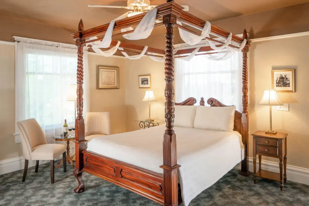 A four-poster wooden bed with white drapes sits in a bedroom featuring two chairs, bedside tables, and framed pictures.