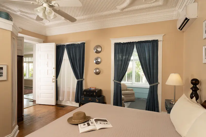 A beige bedroom features a bed with a hat and book, dark blue curtains, and a ceiling fan.