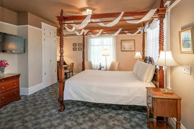A hotel room featuring a large four-poster bed with white drapes, a wooden desk, and a television on the wall.