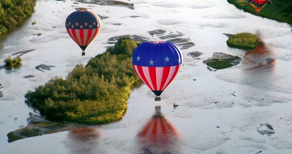 Hot air balloons with stars and stripes designs float over a river and forested islands.