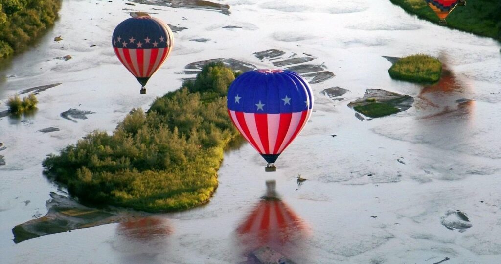 Two American flag-themed hot air balloons fly over a river with grassy islands.