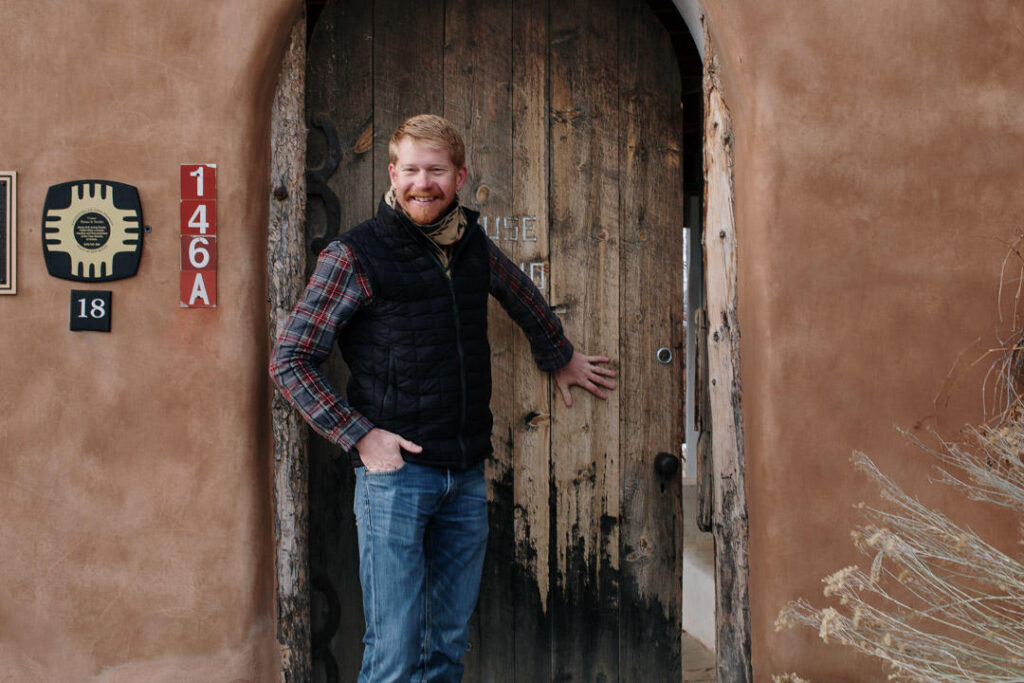A man with a red beard stands outside an arched wooden door in a brown stucco wall, wearing a plaid shirt and vest.