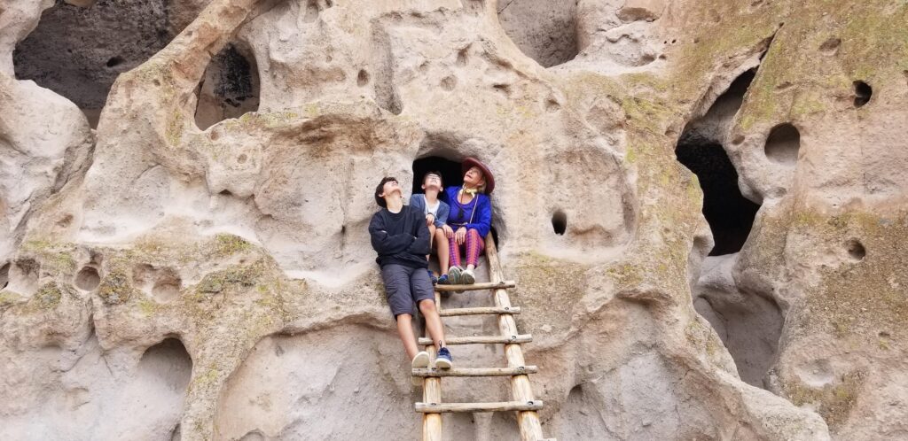 Three people sit on a wooden ladder leading into a small cave entrance carved into a large, textured rock face.