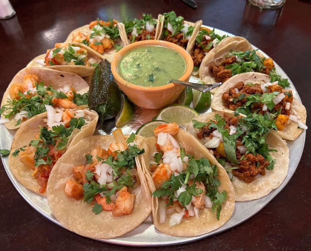 A round platter filled with various street-style tacos arranged around a central bowl of green salsa and lime wedges.