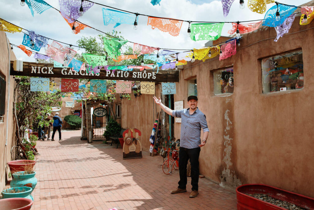 A man gestures toward the courtyard entrance of "Nick Garcia Patio Shops" decorated with colorful Mexican papel picado.