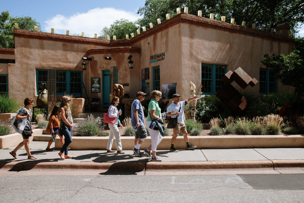 People walk along a sidewalk past a Southwestern-style building with adobe walls and blue-framed windows.