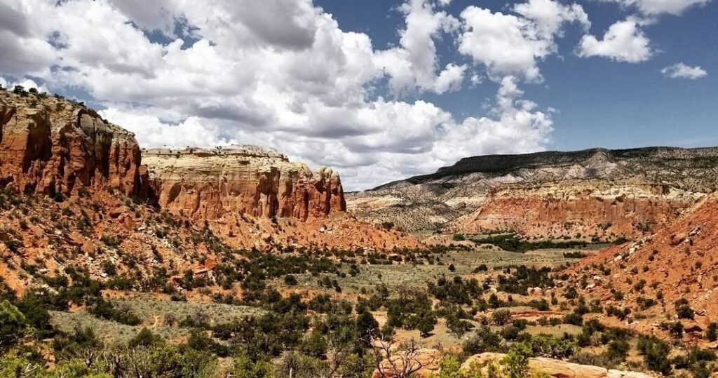 A landscape with red rock formations, sparse vegetation, and a blue sky filled with puffy white clouds.