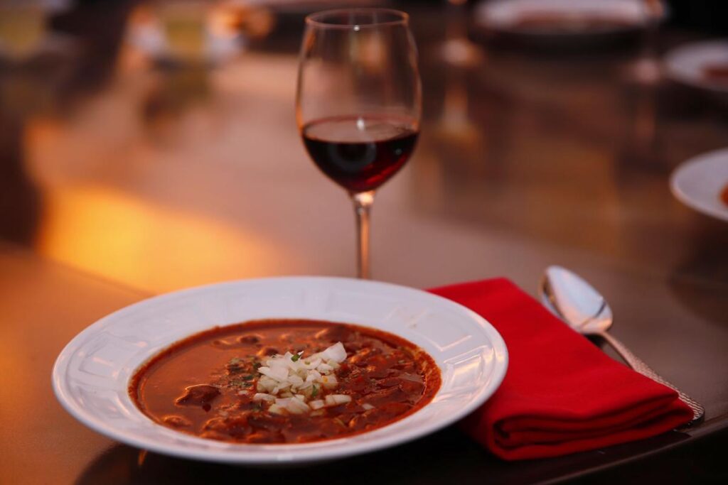 A bowl of red soup with garnish, a glass of red wine, and a red napkin set on a table.