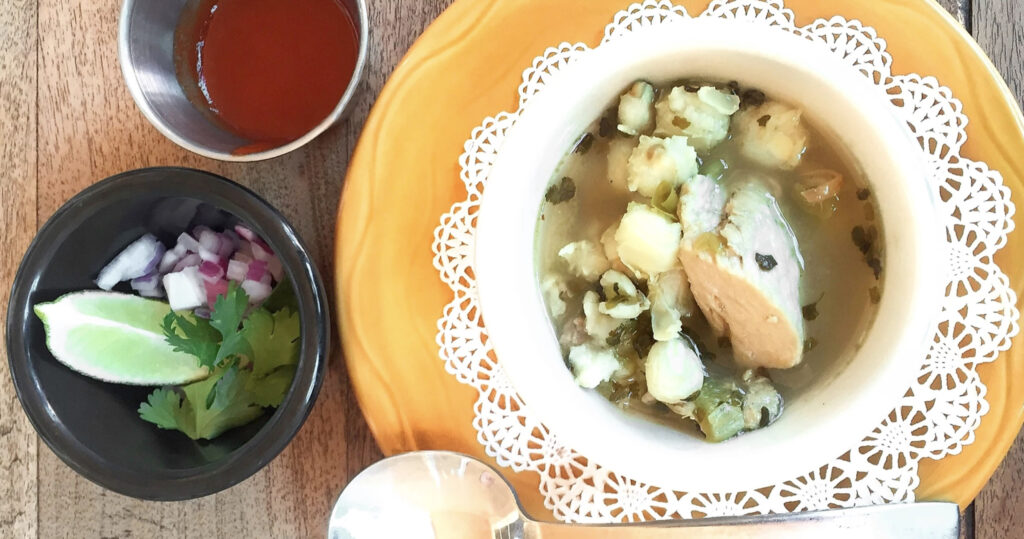 A top-down view of a bowl of seafood soup, sides of lime, onions, cilantro, and chili sauce on a wooden table.