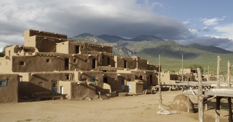 A multi-story adobe structure, resembling the Taos Pueblo, sits in a desert landscape against a backdrop of mountains.