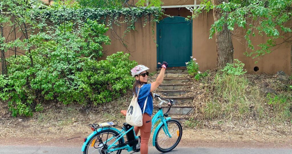 A woman on a turquoise electric bike with a tote bag raises one hand in front of a green gate and a wall.