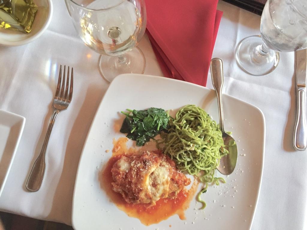 A square plate with a saucy cutlet, pesto pasta, and greens, next to wine and water glasses on a white tablecloth.