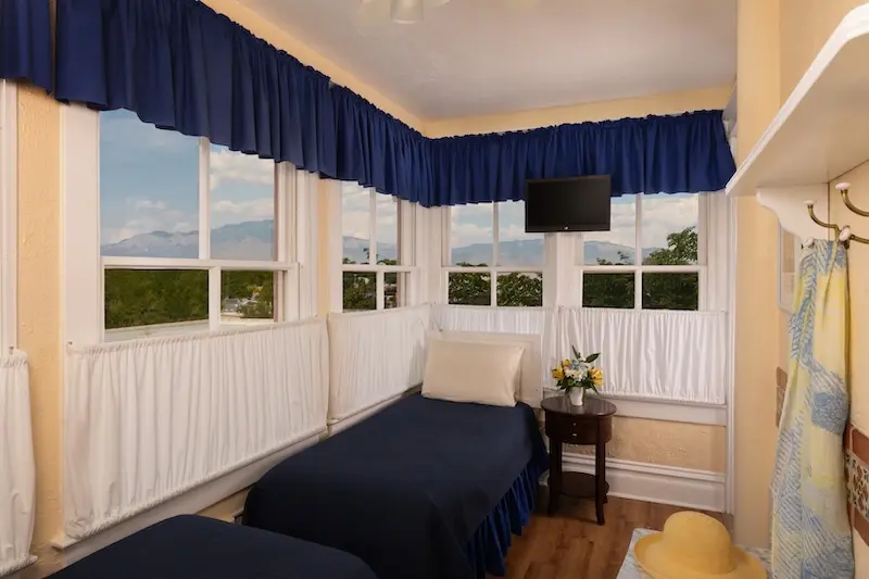 A hotel room with twin beds, blue valances, and windows looking out onto a mountain view.