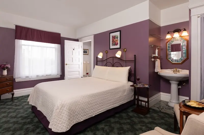 A hotel room with purple walls, a bed, a pedestal sink, a window with curtains, and patterned green carpet.