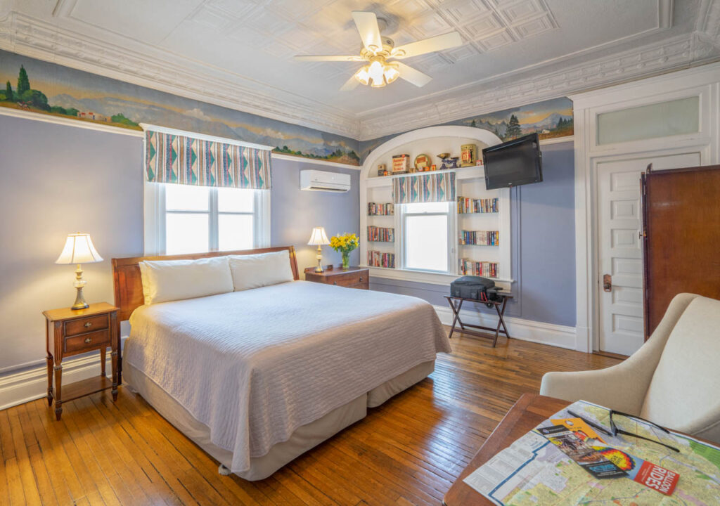 A bedroom with a queen bed, wooden floors, built-in bookshelves, and a ceiling fan in a blue room with a mural.