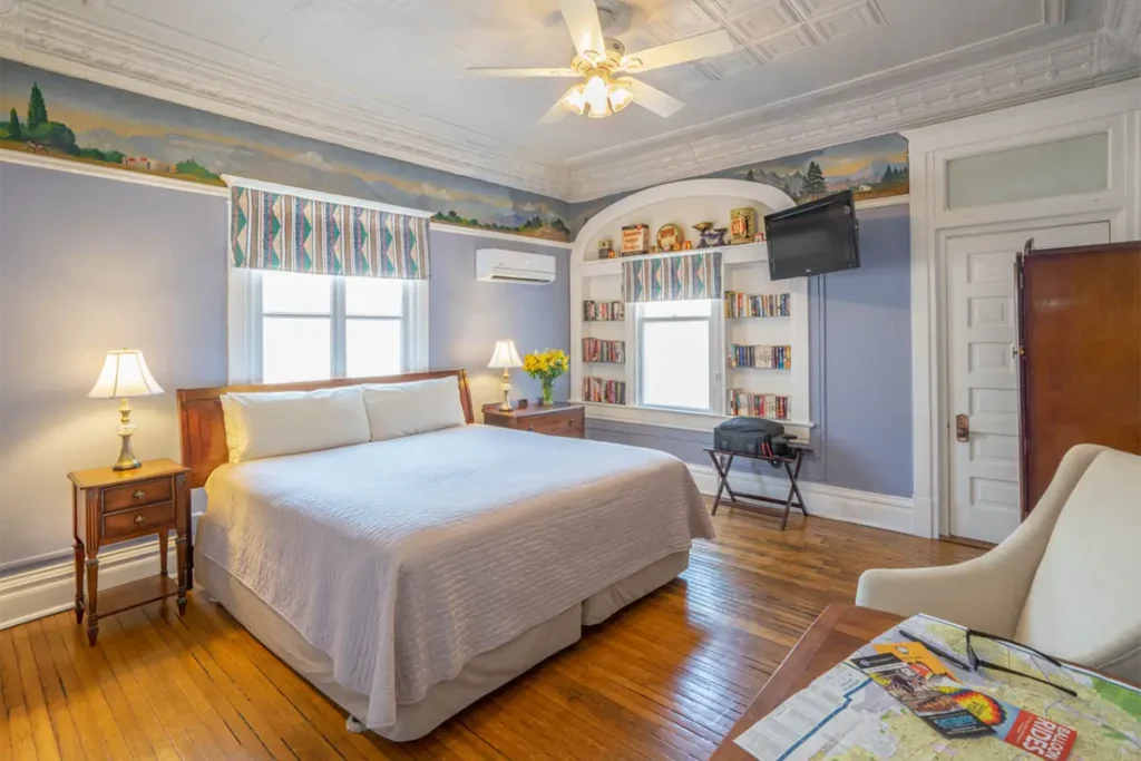 A bedroom features a large bed, hardwood floors, a mural-decorated ceiling, built-in shelving, and a wall-mounted TV.
