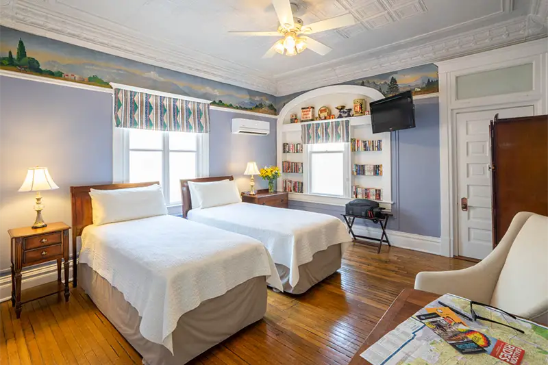 A brightly lit hotel room features two twin beds, hardwood floors, a landscape mural, and a bookshelf with a TV.