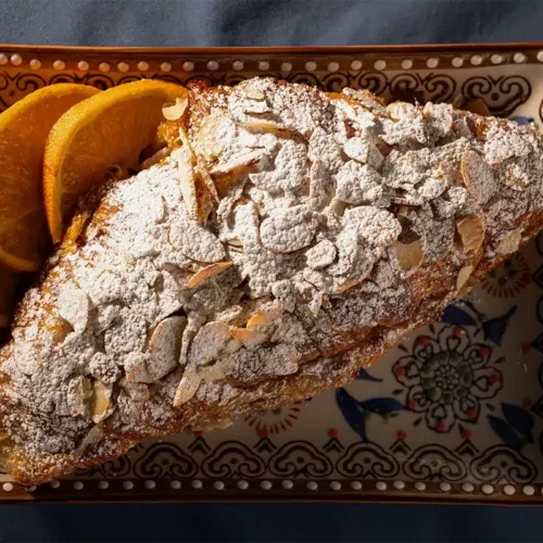 An almond croissant topped with powdered sugar and served with orange slices on a patterned rectangular plate.