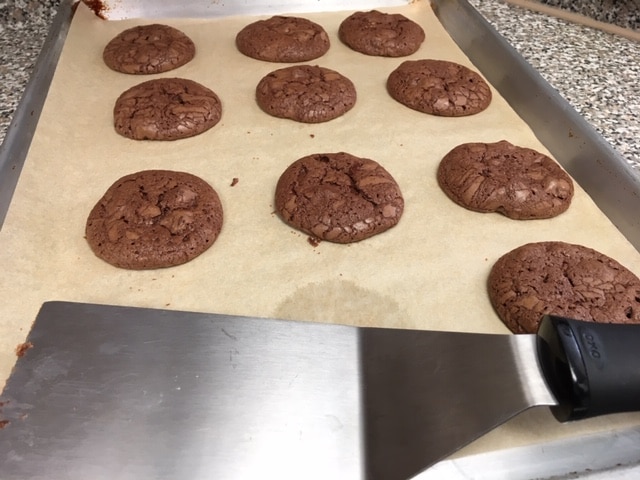 Nine round chocolate cookies lie on a parchment-lined baking sheet next to a metal spatula.