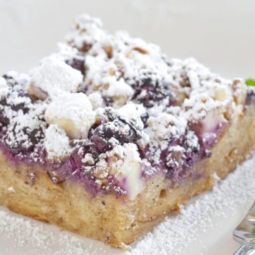 A square piece of blueberry crumble cake dusted with powdered sugar and served on a plate with a fork and mint sprig.