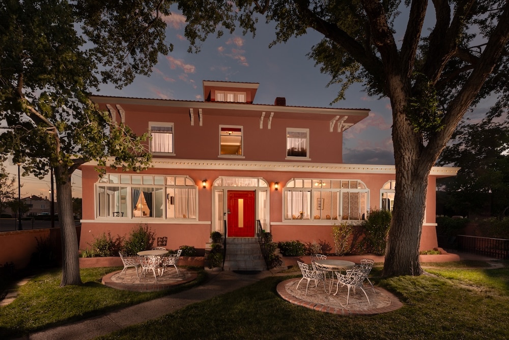 A two-story peach-colored house with a red front door, surrounded by trees and lawn, at twilight.