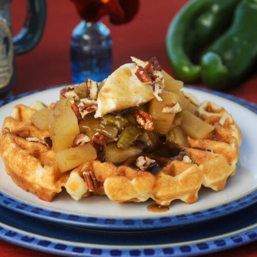 A waffle topped with apples, pecans, and cream on a blue-rimmed plate, with green chiles in the background.