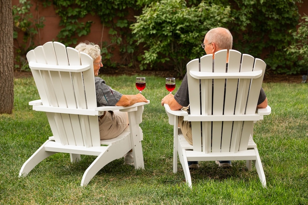 Two seniors sit in white Adirondack chairs on a lawn, holding glasses of red wine and facing each other.