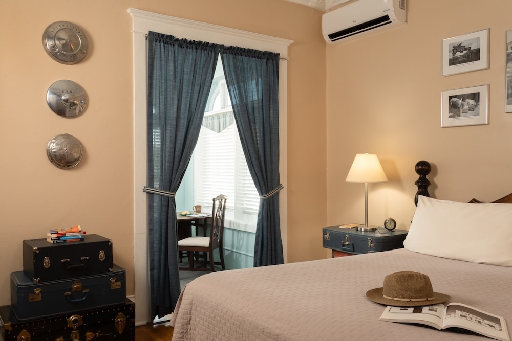A bedroom with a bed, hat, book, vintage trunks, and a window with blue curtains overlooking a small seating area.