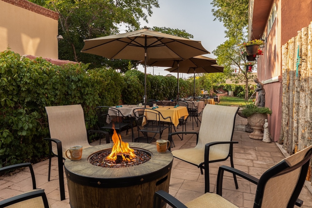Outdoor patio with a fire pit, several chairs, and tables under umbrellas, set against a hedge and brick building.