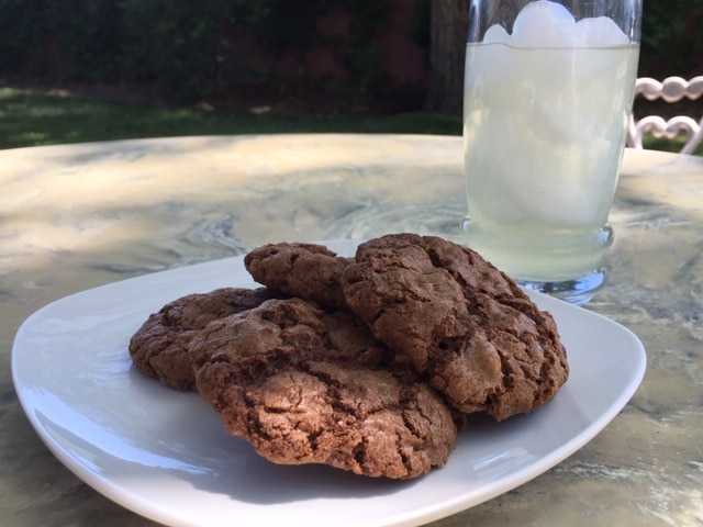 A plate with several chocolate cookies sitting on a marbled outdoor table next to a glass of iced lemonade.