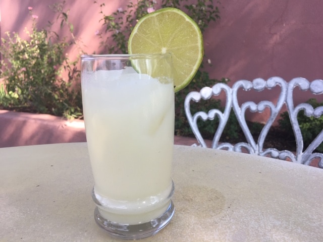 A glass of iced, pale yellow drink garnished with a lime slice, sitting on a table in front of a decorative chair.