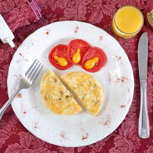A plate featuring a sliced egg frittata and tomato slices sits on a red tablecloth with a fork, knife, spoon, and juice.