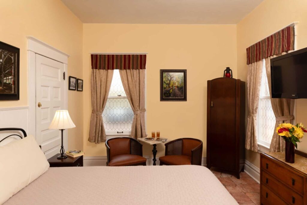 A bedroom with a bed, two chairs, a small table, wardrobe, dresser, TV, and cream walls.