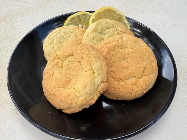 A black plate holds several round, golden-brown cookies garnished with two fresh lemon slices.