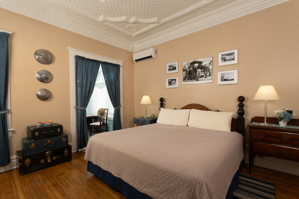 A bedroom features a bed with a beige quilt, two nightstands, framed photos, blue curtains, and a stack of trunks.