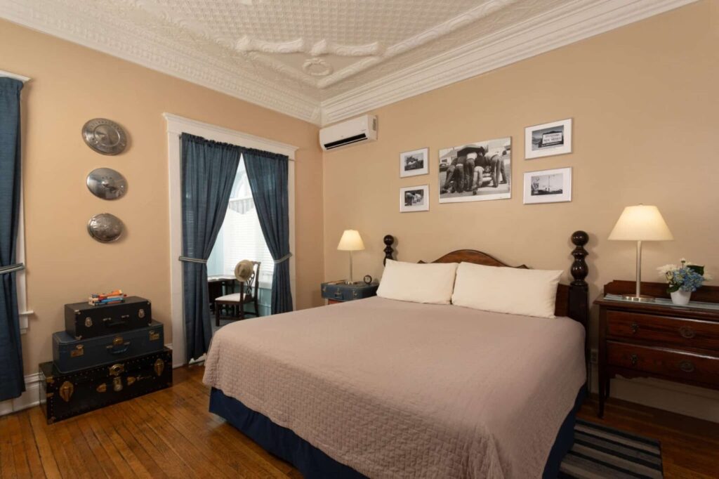 A bedroom with a king bed, beige walls, vintage suitcases, blue curtains, and framed photos on the wall.