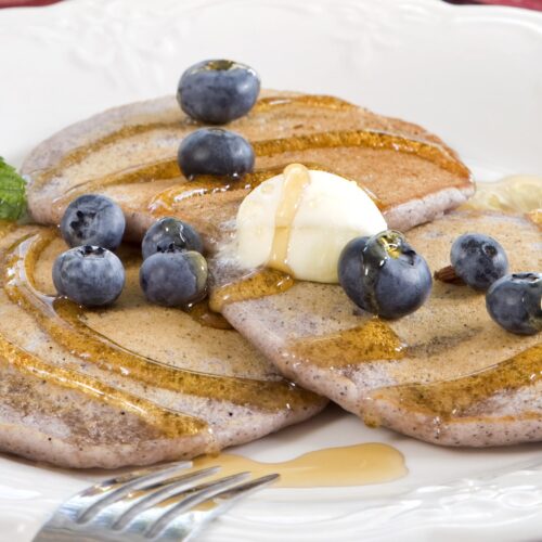 Blueberry pancakes on a white plate are topped with a pat of butter, syrup, and fresh blueberries with a sprig of mint.
