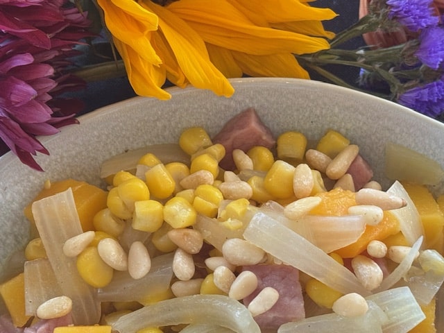 A bowl containing a mixture of corn, diced ham, onions, and pine nuts, arranged below vibrant purple and yellow flowers.
