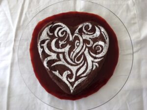 A heart-shaped chocolate cake with a powdered sugar stencil design sits on a glass plate over red sauce.