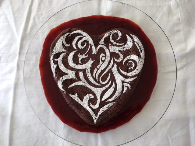 A heart-shaped chocolate cake with a powdered sugar stencil design sits on a glass plate over red sauce.