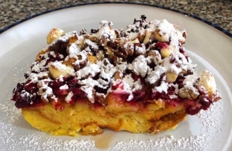 A slice of berry and nut bread pudding topped with powdered sugar rests on a white plate.