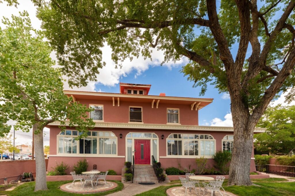 A two-story, reddish-brown house with a red front door, large windows, and a cupola, flanked by two large trees.