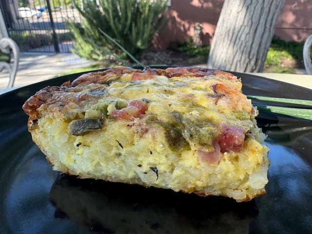 A slice of vegetable quiche on a black plate, set outdoors with blurred greenery in the background.