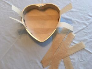 A heart-shaped cake pan is lined with parchment paper strips on a pale surface.