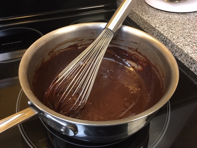 A metal whisk sits in a stainless steel saucepan filled with melted chocolate on a stovetop.