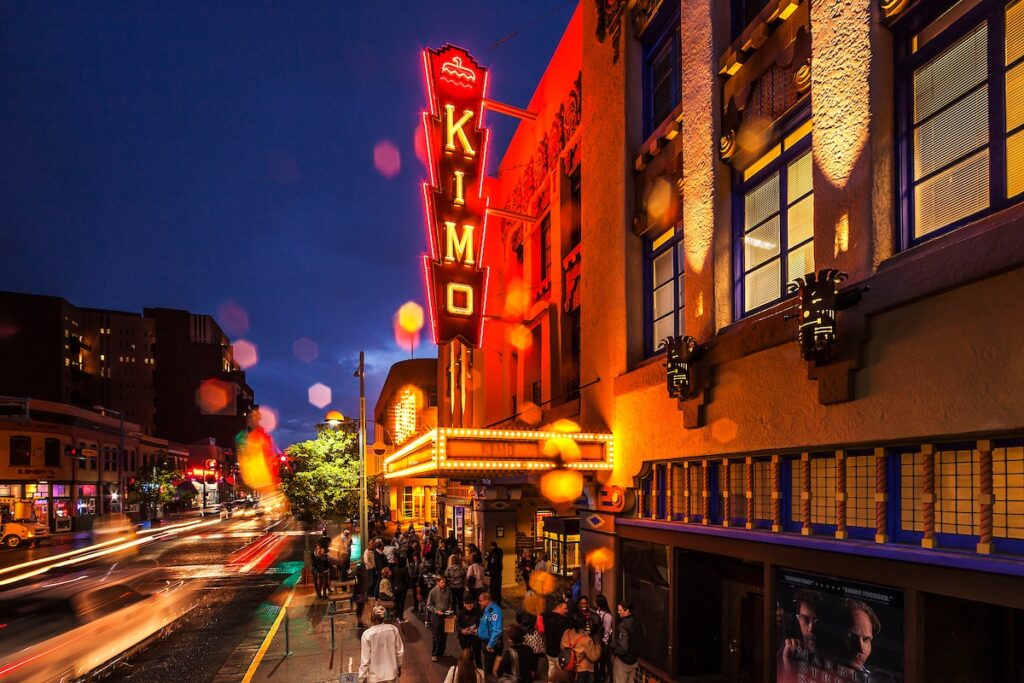 The Kimo Theatre in Albuquerque at twilight, with a glowing red neon sign, street lights, and people on the sidewalk.