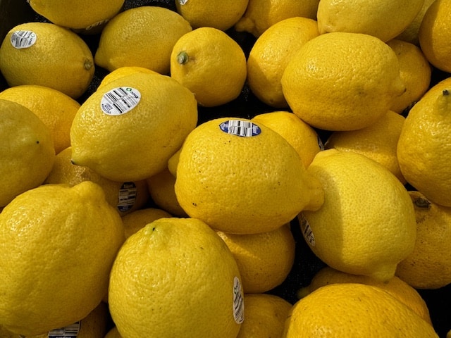 A close-up, high-angle shot of a large pile of bright yellow, textured lemons, some with small white stickers attached.