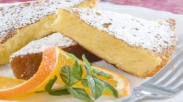 A slice of powdered sugar-dusted cake on a plate with orange slices, a mint sprig, and a fork.