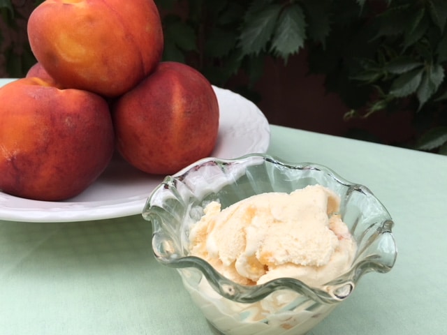 A glass bowl of peach ice cream sits next to a white plate piled with fresh peaches.